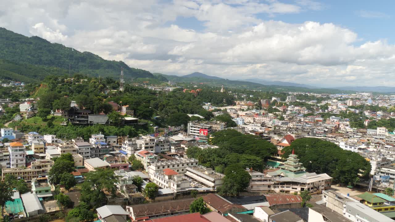 Aerial view of Tachileik town in Shan State, Myanmar (Burma), showing a mix of urban buildings, temples, and lush green hills under a cloudy sky near the Thailand border