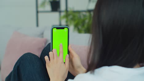 vista trasera de una mujer en una habitación moderna sentada en el sofá usando un teléfono con pantalla verde maqueta de clave de croma con puntos de seguimiento navegando por internet viendo contenido.