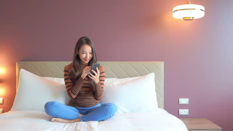 Una-Mujer-Joven-Se-Sienta-En-La-Cama-De-Su-Suite-De-Hotel-Mientras-Envía-Mensajes-De-Texto-A-Su-Teléfono-Móvil