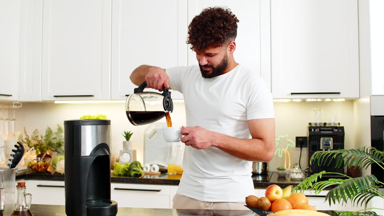 Giovane uomo arabo che versa caffè appena fatto nella cucina di casa, al mattino, dipendenza quotidiana da caffeina