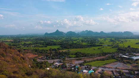 a view from a peak in thailand overlooking green rice fields and towering mountains in the distance surrounded by a colorful forest