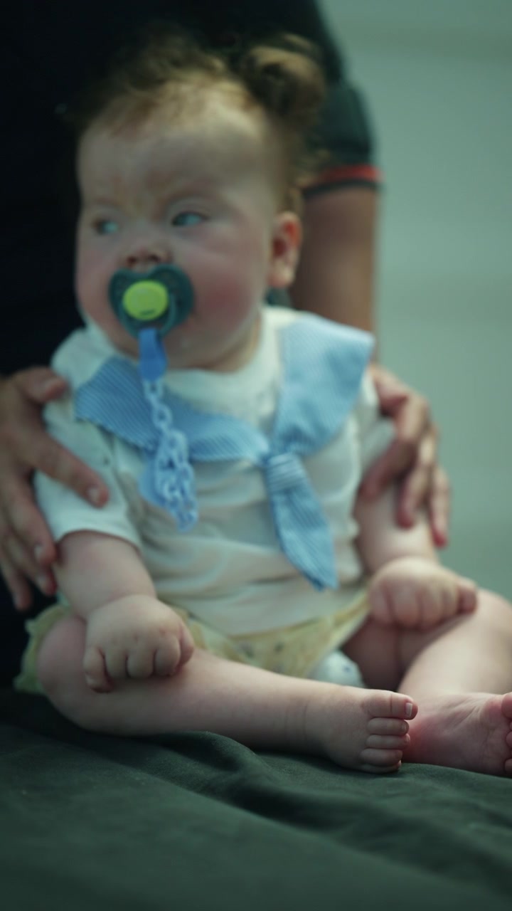 Baby boy holding pacifier, looking directly into the camera. An adult's hands are gently supporting the infant