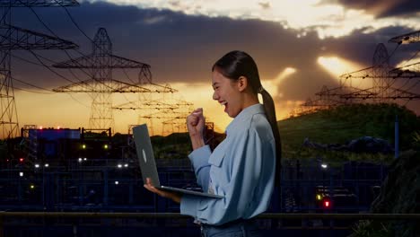 vista lateral de una trabajadora profesional asiática de pie con su portátil cerca de la torre de alto voltaje, instalación industrial, ella levanta el puño con una meta gritando después de escribir en la computadora portátil