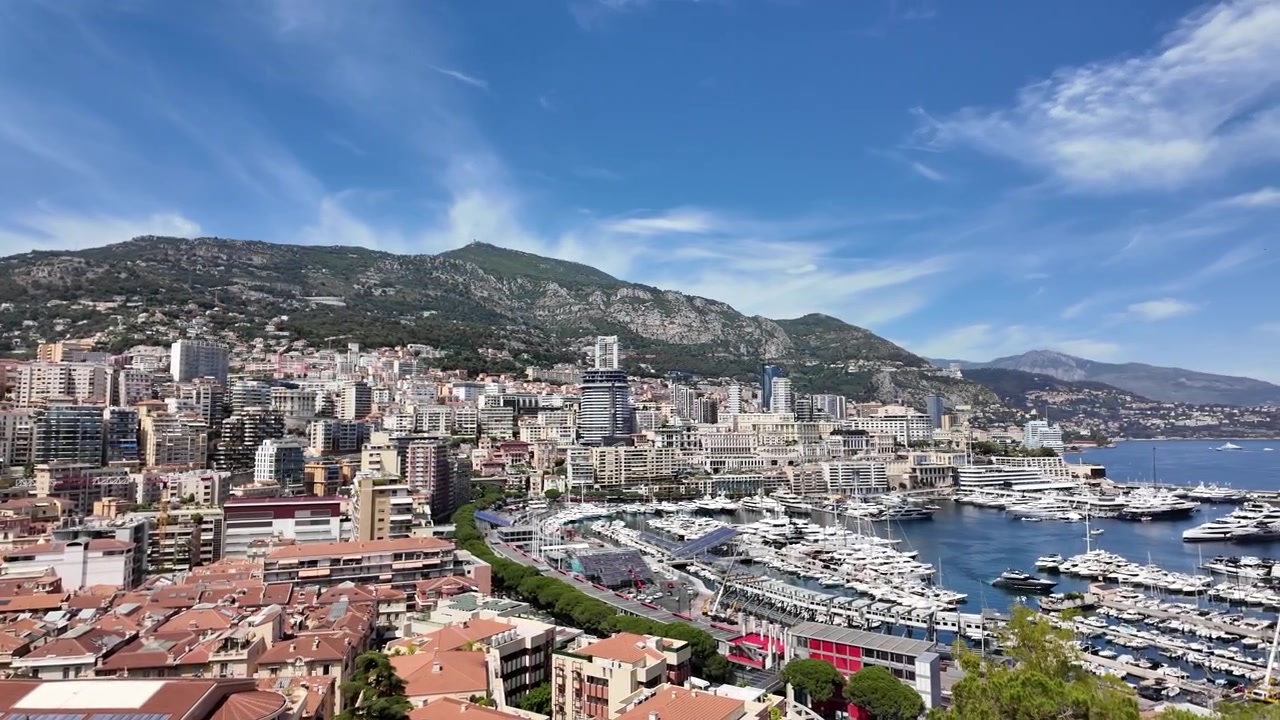 Vista panoramica di Monaco e Port Hercule con yacht di lusso, skyline moderno e costa mediterranea incorniciata da montagne imponenti sotto un cielo azzurro brillante in una giornata di sole