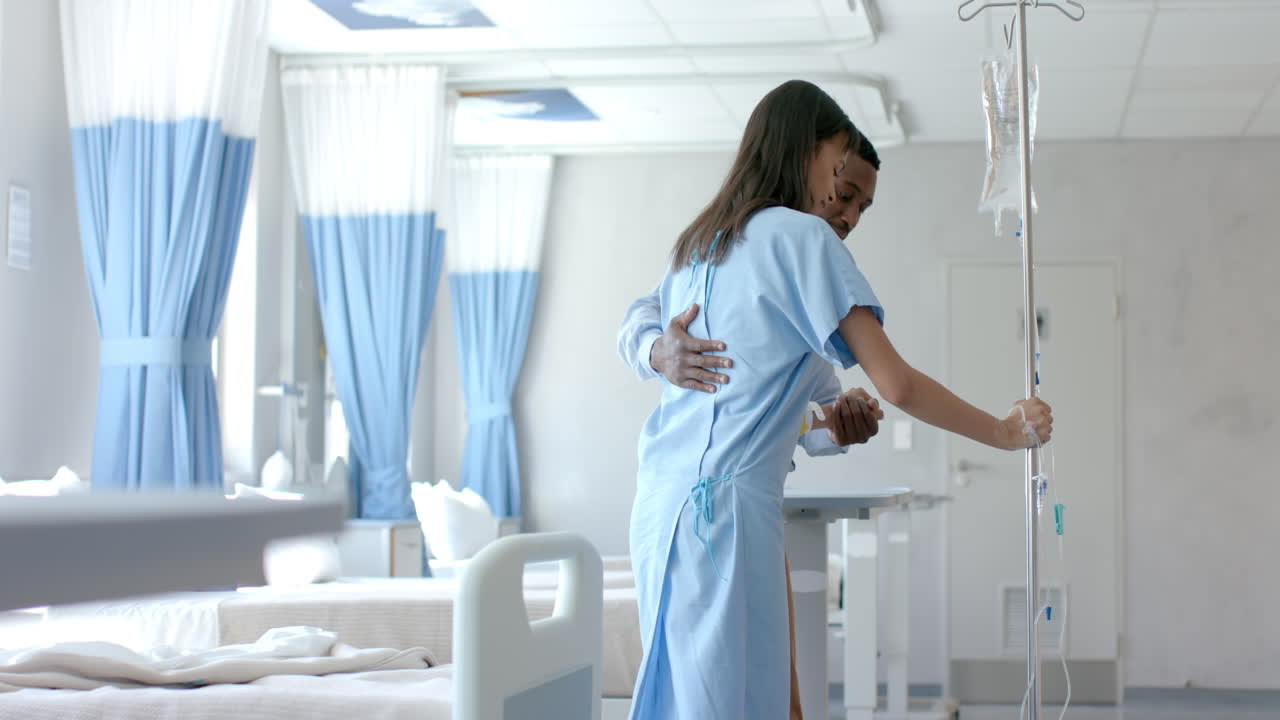 médico varón diverso ayudando a una paciente a caminar con un hilo de goteo en la sala del hospital, en cámara lenta