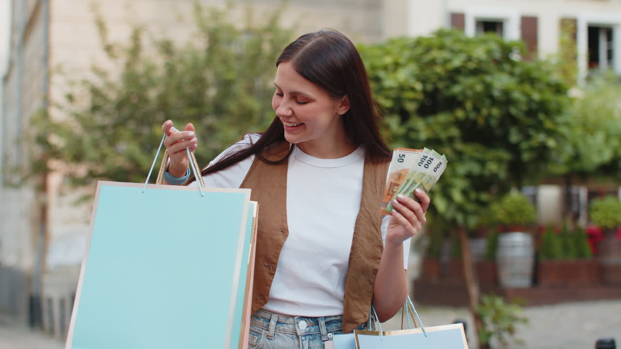 joven adicta a las compras mujer caucásica feliz sosteniendo efectivo en euros mientras lleva bolsas de compras en la calle de la ciudad
