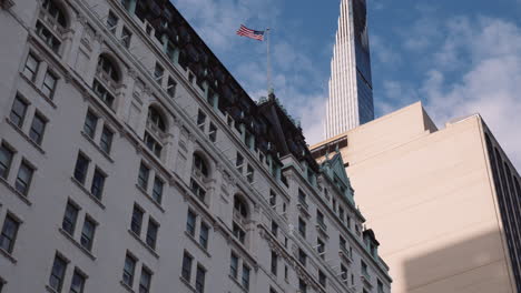 low angle view of plaza hotel building facade in new york city manhattan usa, american flag on top of building, urban architecture and modern downtown tower in background