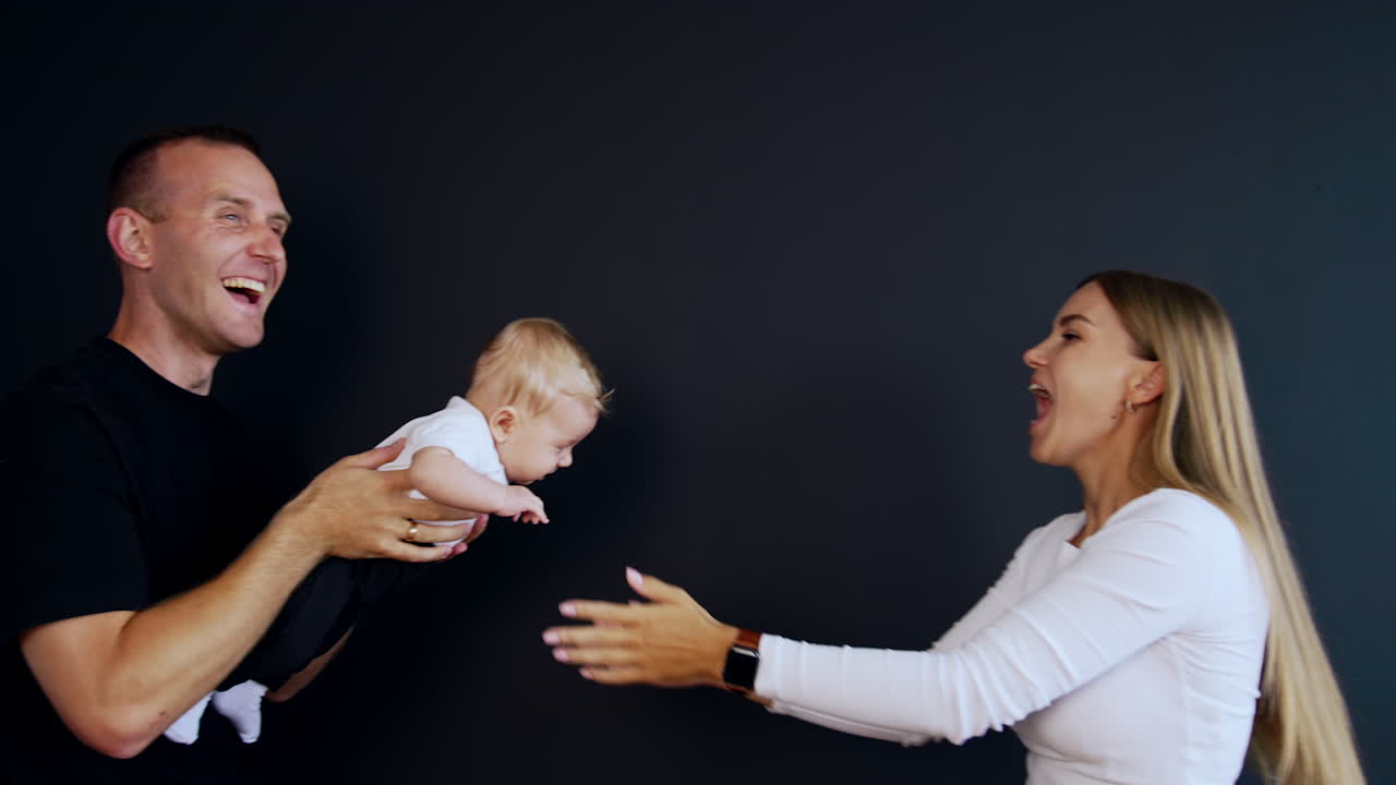 Happy Caucasian parents playing with their adorable infant baby. Lovely family of three having fun. Black backdrop.