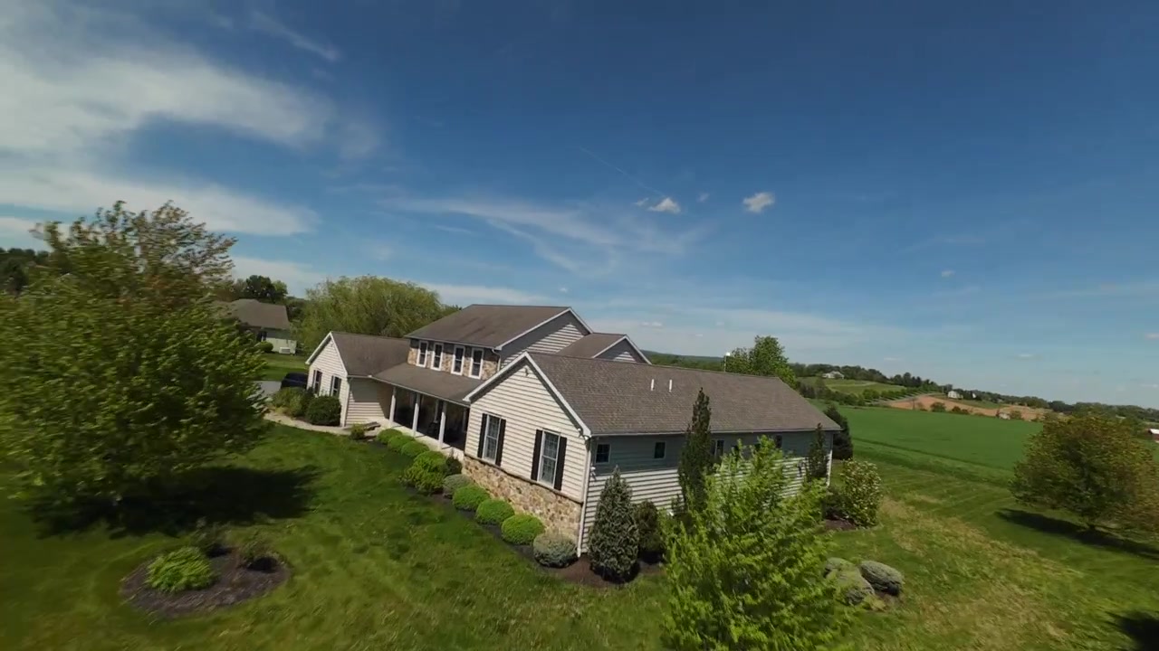 Vol FPV au-dessus d'une maison individuelle avec des champs verts et une ferme en arrière-plan. Journée ensoleillée avec ciel bleu au printemps. Survol dynamique.