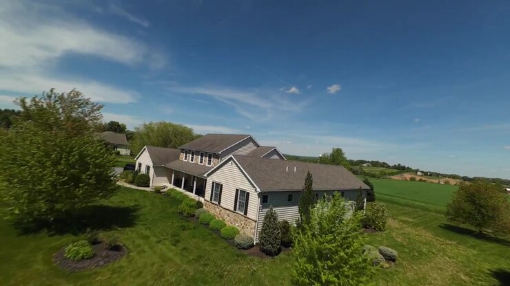 Vol FPV au-dessus d'une maison individuelle avec des champs verts et une ferme en arrière-plan. Journée ensoleillée avec ciel bleu au printemps. Survol dynamique.