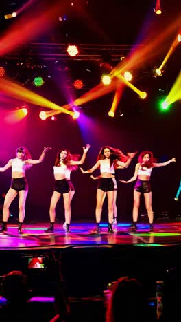 four k-pop idols performing an energetic dance routine on a brightly lit stage, captivating an enthusiastic audience while fans eagerly recording the vibrant show with smartphones