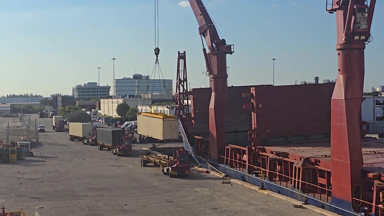 A massive red cargo ship is docked at a busy port terminal during daytime. Cranes and trucks operate on the concrete dock, handling shipping containers and freight for global trade and logistics.