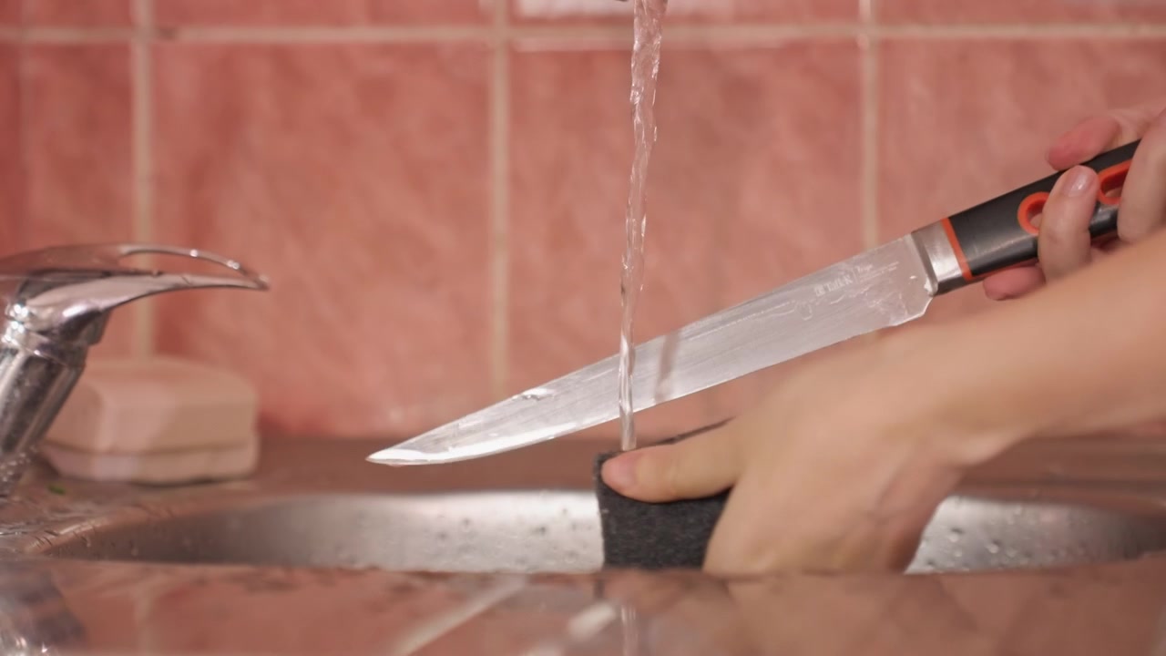 Primo piano di mani che lavano un coltello sotto l'acqua corrente. Le mani strofinano l'utensile metallico con una spugna, concentrandosi. Un lavello in acciaio inossidabile è visibile per tutta la scena, enfatizzando l'utensile.