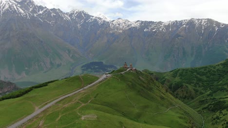 drone aerial view in georgia flying towards gergeti trinity orthodox church in kazbegi surrounded by green mountains valley with snowed peaks