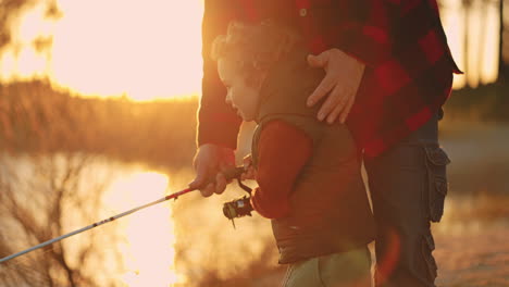 little boy anf his father are fishing together on river or lake shore in sunset time