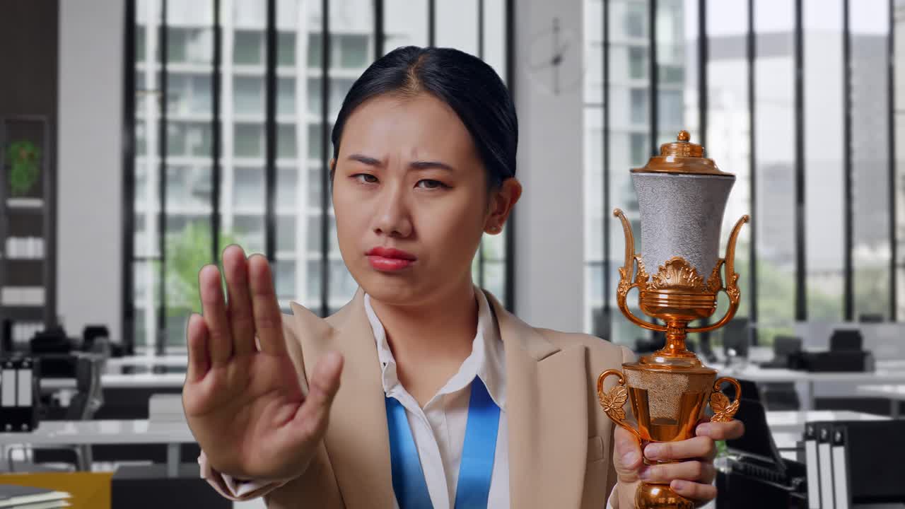Close Up Of Asian Business Woman In A Suit With A Gold Medal And Trophy Disapproving With No Hand Sign And Smiling To Camera In The Office