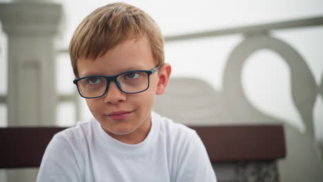 a little boy wearing glasses sits alone with a serious expression, appearing to hold back a smile as he adjust on the chair