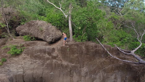 caucasian man in blue swim shorts walks along edge of steep rock cliff