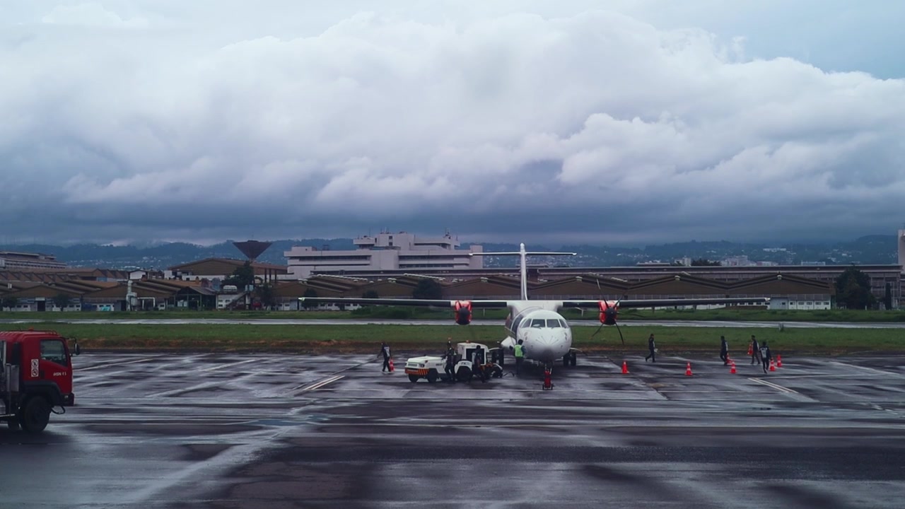 濡れた空港の滑走路に駐機し、近くで地上作業員が作業する小型プロペラ機。