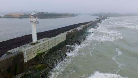 vista aérea del muelle de hormigón del puerto de liepaja, costa del mar báltico, día brumoso con niebla, sensación de mal humor, grandes olas de tormenta salpicando, faro blanco, disparo de drones avanzando