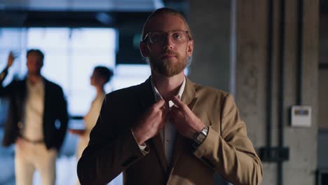 Portrait-of-a-blond-man-with-a-beard-who-adjusts-his-light-brown-suit,-folds-his-arms-on-his-chest,-smiles-and-looks-at-the-camera-against-of-a-modern-office