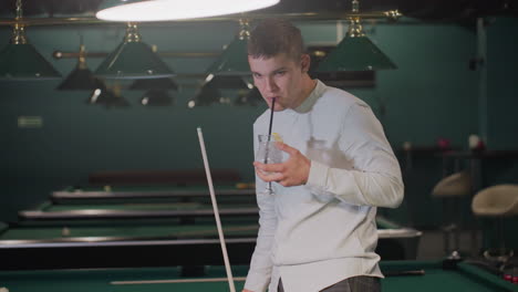 Billiard player in white shirt and grey trousers holds cue stick and sips lemon drink while pacing in dimly lit pool hall. Green pool tables and overhead lamps create an atmospheric setting