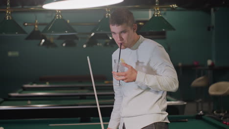 billiard player in white shirt and grey trousers holds cue stick and sips lemon drink while pacing in dimly lit pool hall. green pool tables and overhead lamps create an atmospheric setting