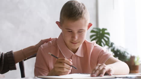 close-up view of a boy with down syndrome doing homeworks sitting at table in the living room at home. his mother supports him and caresses his shoulder