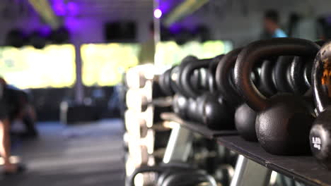 a rack of weights and kettlebells in a gym with people defocused working out and training in the background