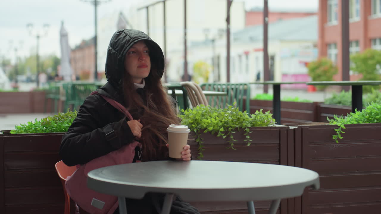 Donna con cappuccio che tiene in mano un caffè, terrazza di un bar all'aperto sotto una pioggia leggera, edifici in mattoni sullo sfondo, borsa rosa a tracolla, sorseggia lentamente con sguardo pensieroso, atmosfera accogliente e malinconica, tonalità cinematografiche