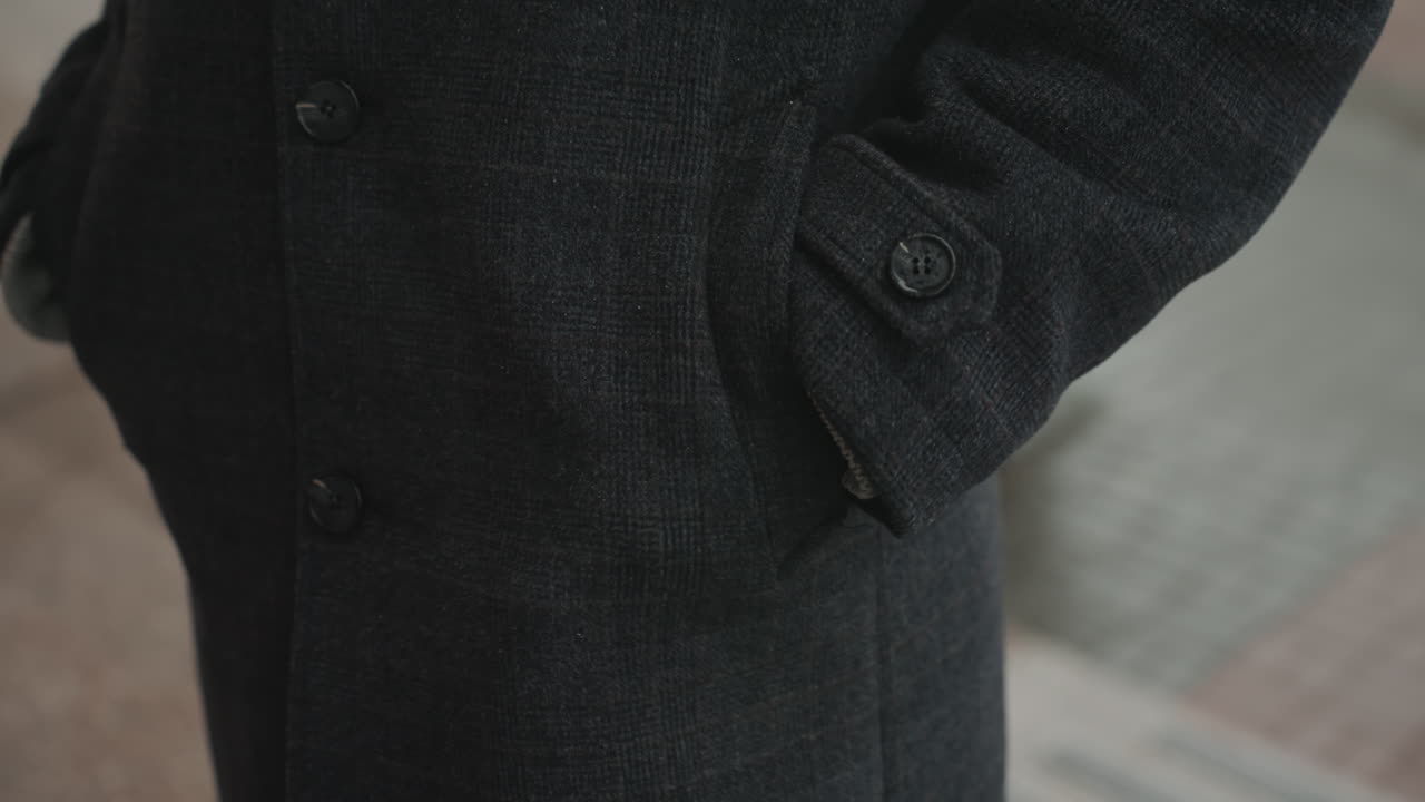 Creatore in cappotto nero in piedi con le mani in tasca, tessuto di lana testurizzato. Primo piano di manica e polsino abbottonati. Ambientazione cinematografica su un marciapiede urbano. Una sottile immobilità suggerisce un creatore riflessivo che si prepara.