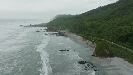 reverse aerial ocean view of wild west coast on the south island of new zealand aotearoa, with waves rolling into shoreline and green, tree covered landscape