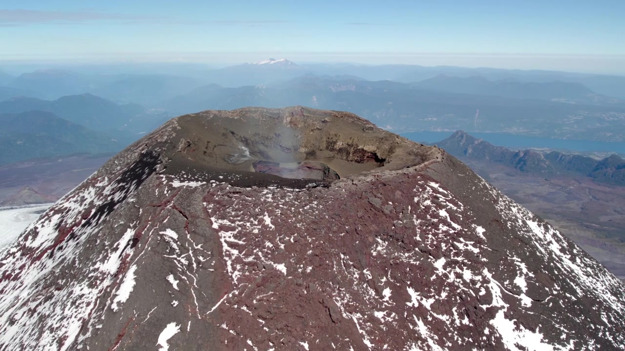 A breathtaking view from the summit of a volcano reveals a large crater surrounded by rocky terrain, with a serene lake visible in the distance against a clear blue sky.