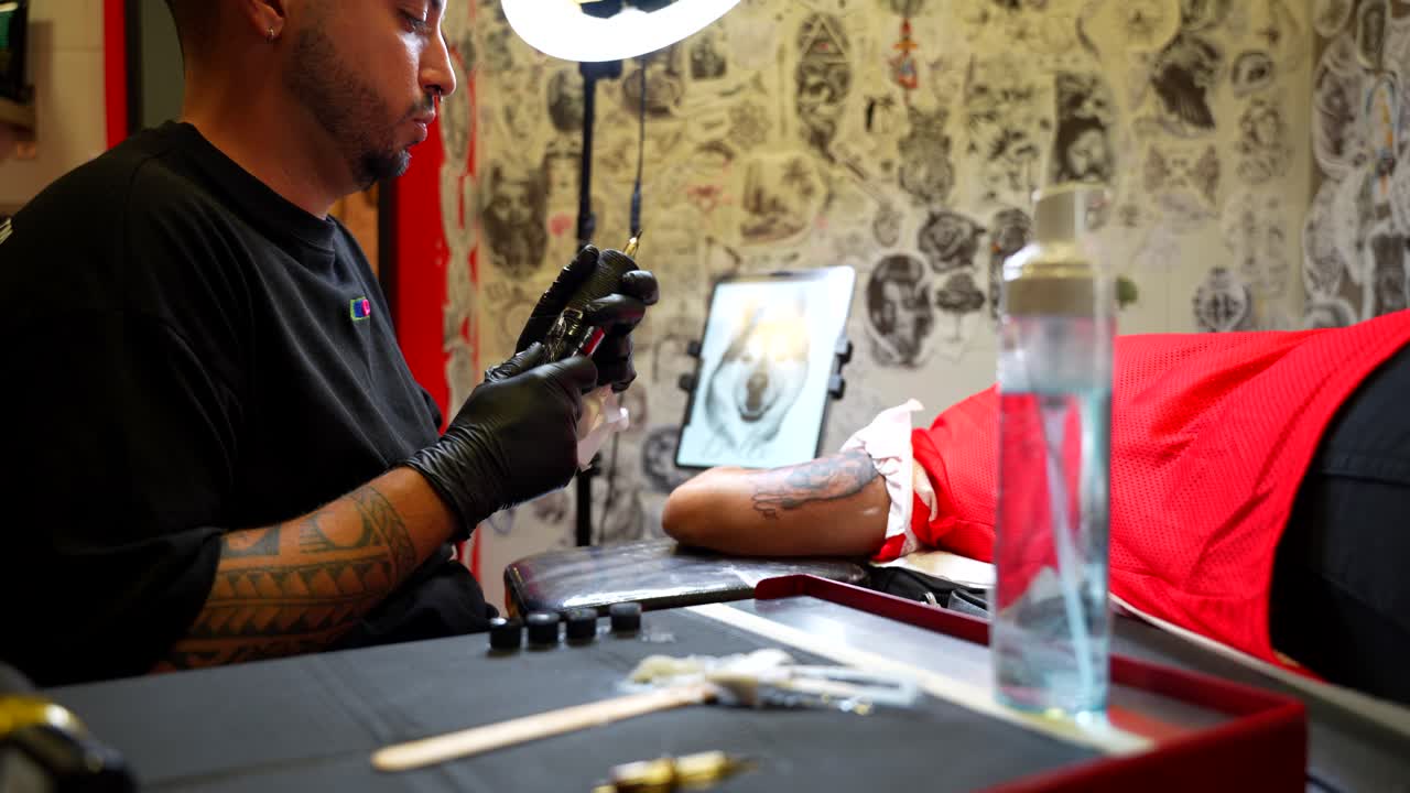 Tattoo artist changes the spike of the pen, takes some ink and continues drawing a dog's face on the arm of his female customer