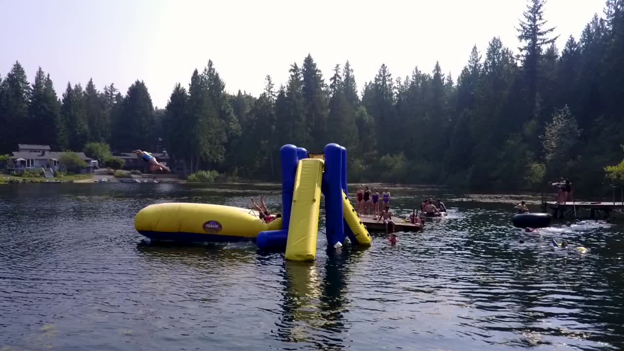 A kid jumps onto an inflatable air cushion and sends their friend flying into the air and then into a lake in a slow motion, drone shot