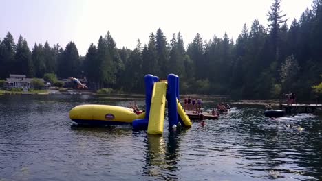 A-kid-jumps-onto-an-inflatable-air-cushion-and-sends-their-friend-flying-into-the-air-and-then-into-a-lake-in-a-slow-motion,-drone-shot