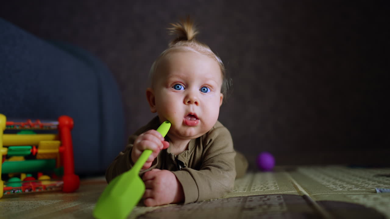 Cute little infant with adorable plump cheeks and blue eyes. Sweet baby lies on the floor chewing a toy shovel. Close up.