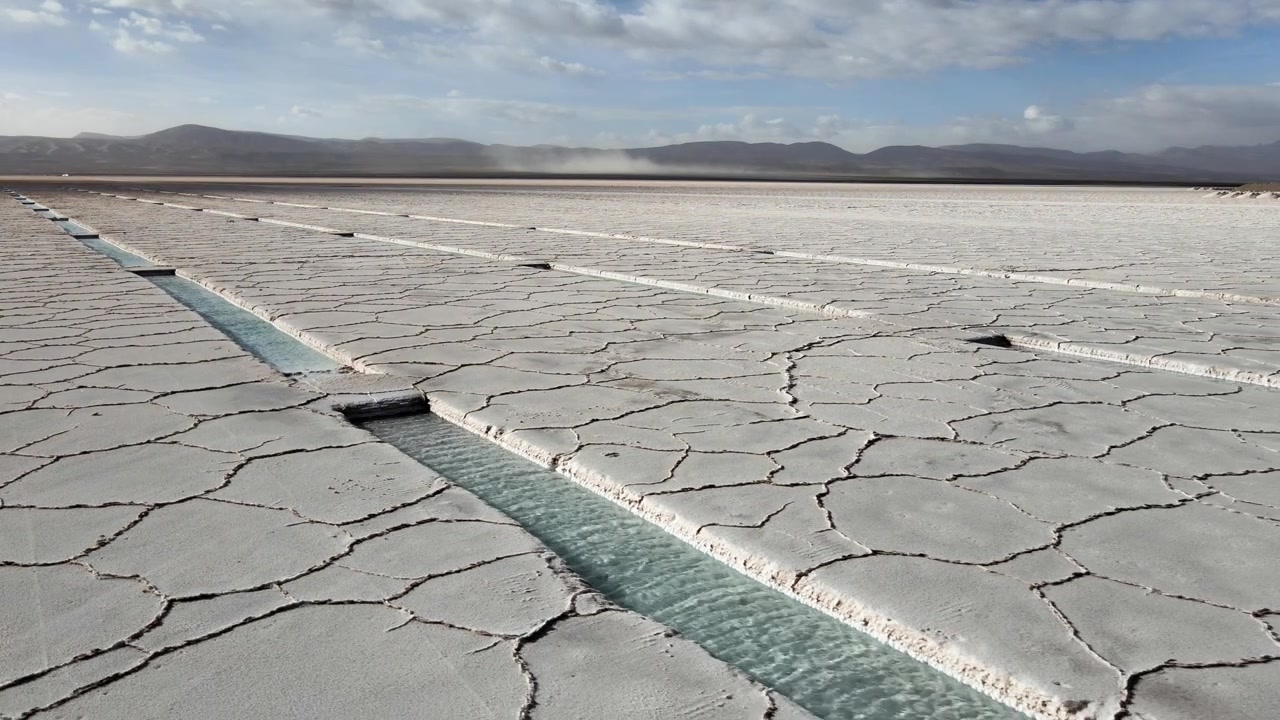 Shallow turquoise lithium brine canals run across the cracked white surface of Salinas Grandes salt flat in Jujuy Province under a bright daytime sky.