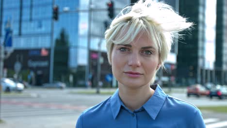 young blonde woman in a big city staring into the lens with a neutral look and hair blowing in the wind