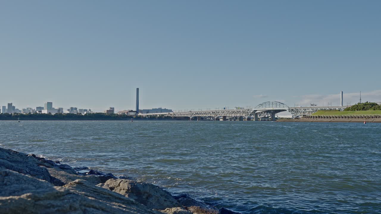 Wide shot of a modern white arch bridge spanning the water, with a rocky foreground and a dense urban skyline in the background