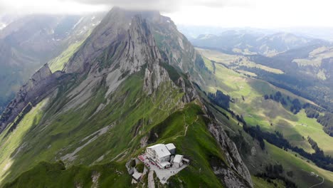 Aerial-flyover-towards-the-cliffs-of-Schafler-ridge-in-Appenzell,-Switzerland-with-mountain-hut,-mountain-peaks-and-lush-green-mountainsides-in-view