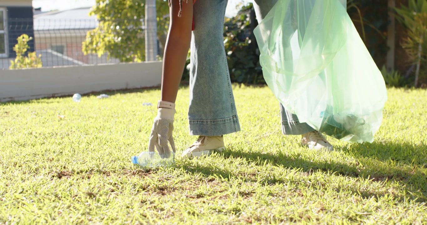 Woman spotting bottle, bending, grabbing while wearing gloves, placing in green bag, cleaning yard