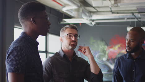 three millennial male creatives stand having a discussion in their workplace, close up