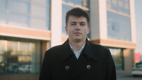 man in black coat stands outdoors with a subtle smile, exuding confidence and poise, blurred background featuring modern building highlights urban professional setting