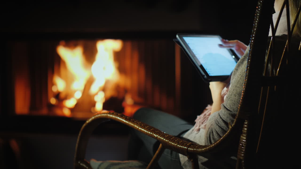 una mujer usa una tableta mientras se sienta en una mecedora junto a la chimenea en casa