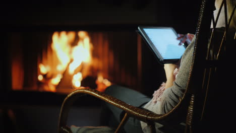 a woman uses a tablet while sitting in a rocking chair by the fireplace at home