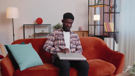 African-American-man-sitting-on-sofa-opening-laptop-pc-starting-work-online-in-living-room-at-home