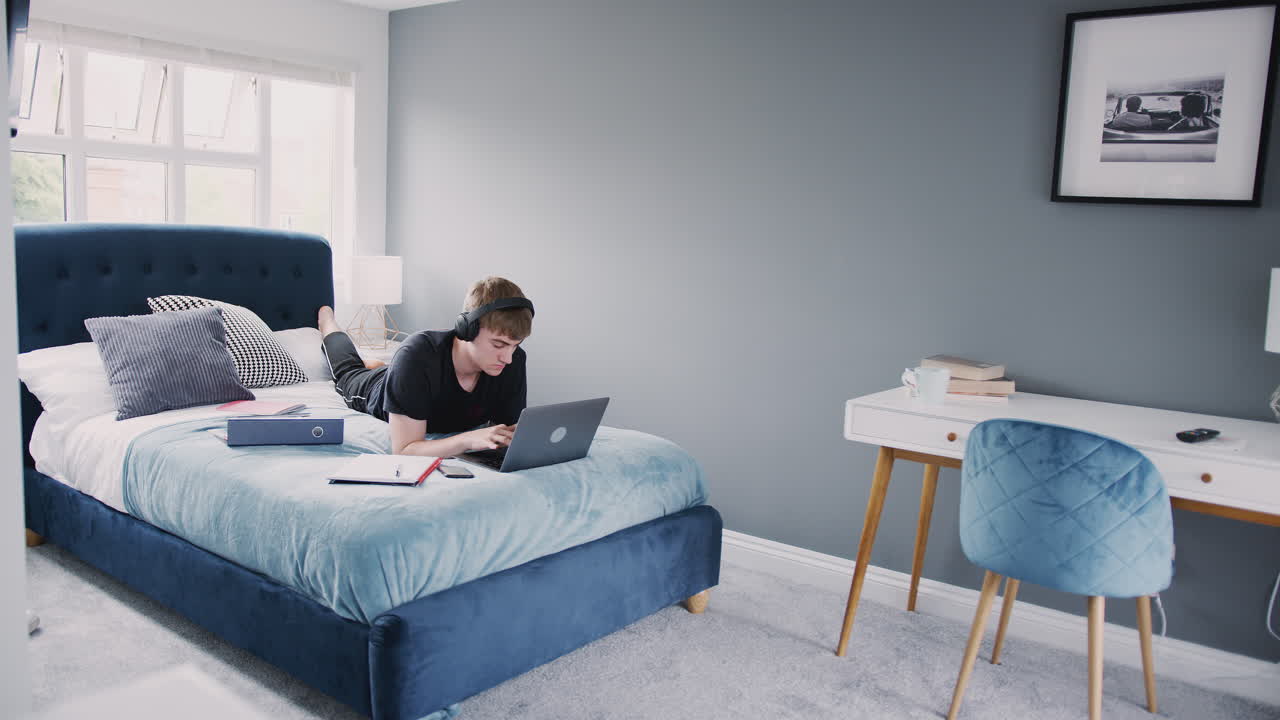 estudiante universitario masculino con auriculares trabaja en la cama en una casa compartida con portátil y teléfono móvil