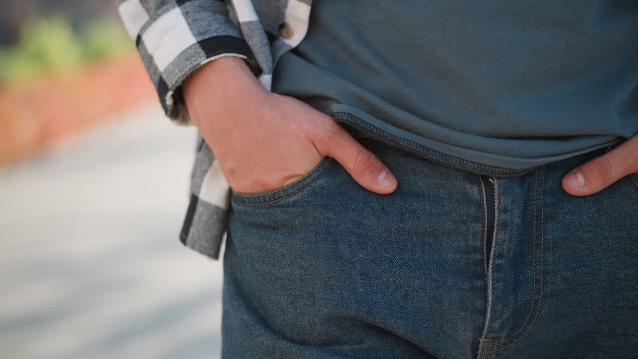 Close up man with hand in jeans pocket walking slowly along sunlit pathway with soft blur background of greenery and urban structures during clear day with long shadows cast