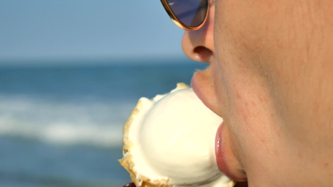 joven mujer morena lamendo comiendo helado en la playa, fondo de mar azul y gafas de sol.